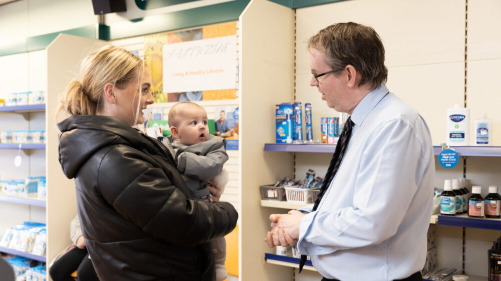Pharmacy worker talking to a person holding a baby in a pharmacy setting