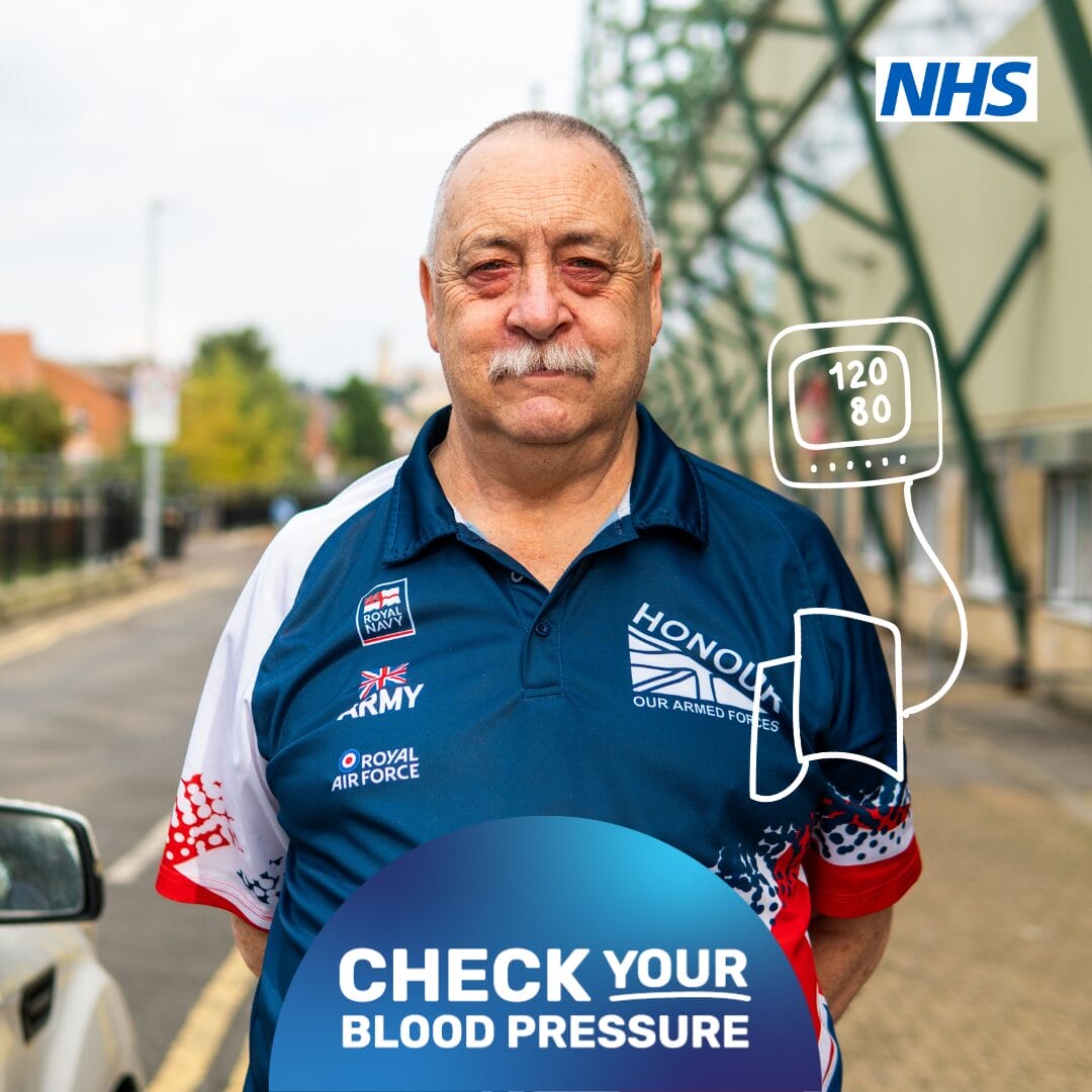 Person standing facing the camera with his hands casually linked behind his back, in a street, smiling. Cartoon on the photo shows a blood pressure monitor on his arm and a reading.