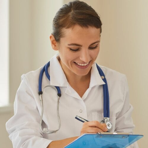 A female Clinician wearing a white coat with a stethoscope around her neck, writing on a clipboard