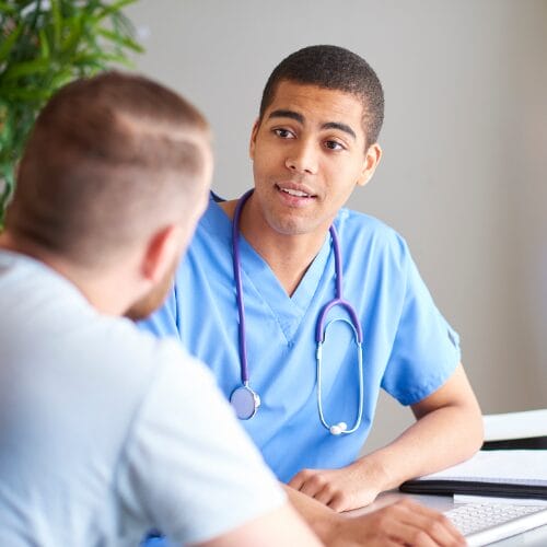 A male Clinician wearing blue scrubs sat at a desk talking to a patient