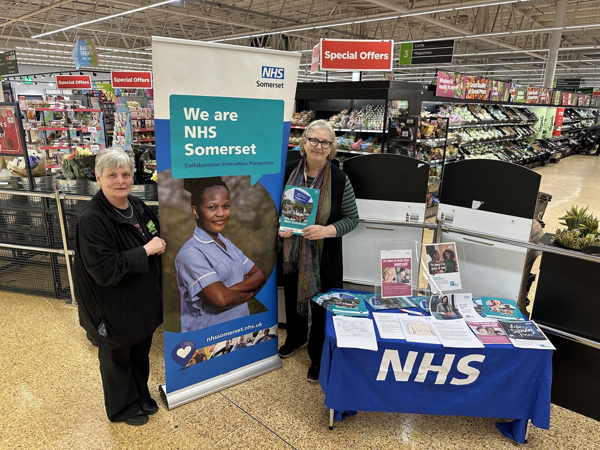 2 people stood next to an NHS Somerset banner and table with documents on in a supermarket.