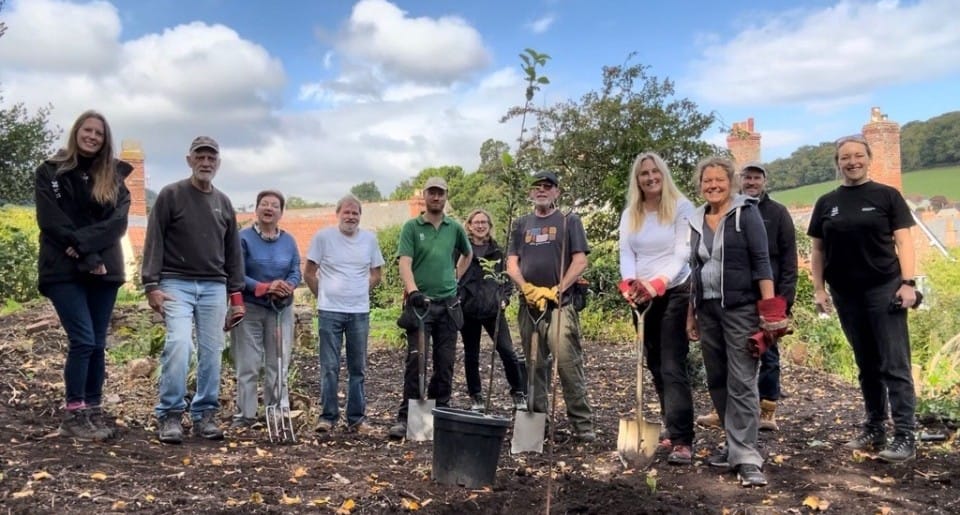 Image shows a group of 11 people, including members of the NHS Somerset Armed Forces team, with spades and forks standing in the grounds of Dunster Castle with a one of the trees they have planted.