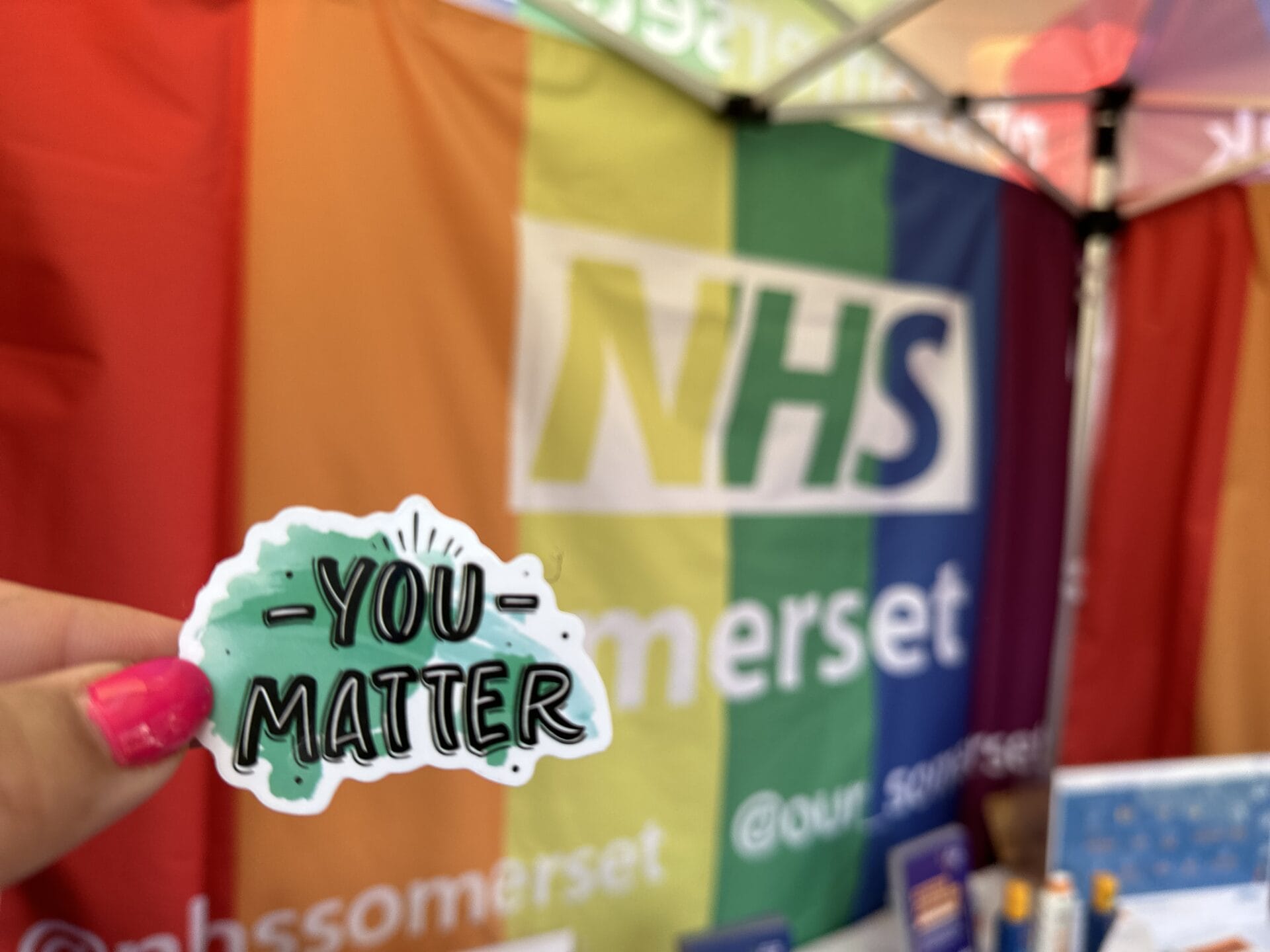 Picture of NHS Somerset's rainbow gazebo with a hand holding a sticker in the forefront.