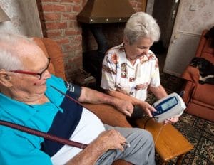 2 people sat in a living room one with a blood pressure cuff on and the other holding the blood pressure monitoring machine.