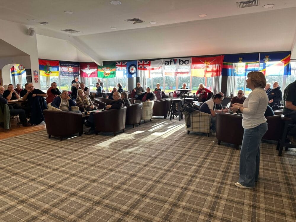 Image is of a room, full of people. Armed Forces Lead for NHS Somerset, Teri Underwood is standing at the front speaking to a group of people.
