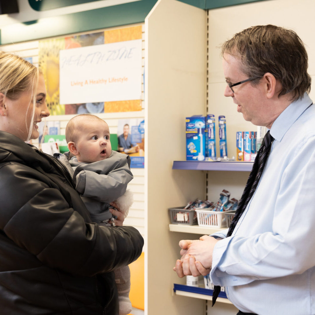 Pharmacy worker talking to a person holding a baby in a pharmacy setting