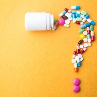 A white plastic tablet bottle positioned horizontally with a variety of tablets and capsules pouring out of it in the shape of a question mark