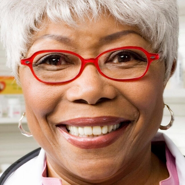 A smiling pharmacist in red glasses standing in front of shelves at a pharmacy